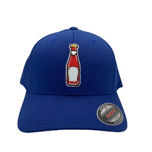 May include: A royal blue baseball cap with a red and white ketchup bottle embroidered on the front. The cap has a black Flexfit label on the back with the text "The Original Flexfit, UK, It Fits Your Style".