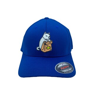 May include: A royal blue baseball cap with a Flexfit design, featuring an embroidered cartoon cat. The cat is shown with a brown paper bag and a fish. The hat has a curved peak and a Flexfit sticker.