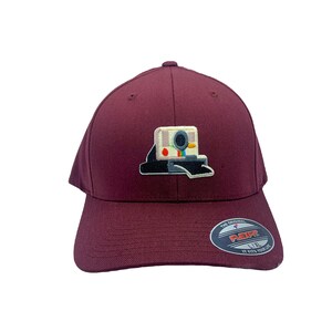 May include: A maroon baseball cap with a white and black camera graphic on the front. The cap has a black Flexfit label on the back with the text "The Original Flexfit L/XL It Fits Your Life."