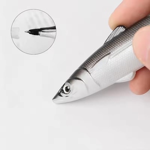 May include: A novelty pen shaped like a fish. The pen has a silver body with gray detailing and a black pen tip. The pen is held by a hand against a white background. A close-up of the pen tip is in a circular inset.