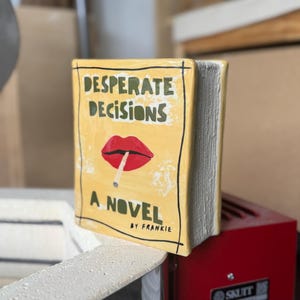 May include: A yellow ceramic book sculpture with a glossy finish. The cover features the text "DESPERATE DECISIONS", "A NOVEL", and "BY FRANKIE" in black, with a red graphic of lips holding a cigarette. The side shows textured white pages.