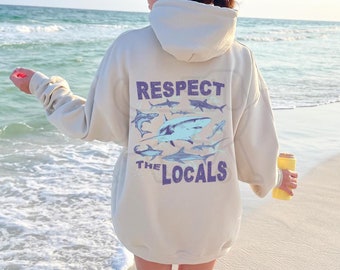 May include: A beige hoodie with a graphic print of sharks and the text "Respect the Locals".