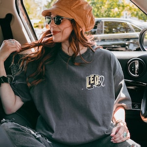 Puede incluir: Una mujer con una camiseta gris con la palabra "LEO" en letras amarillas en la parte delantera. Est&aacute; sentada en el asiento del conductor de un coche y lleva un sombrero de pana marr&oacute;n y gafas de sol.