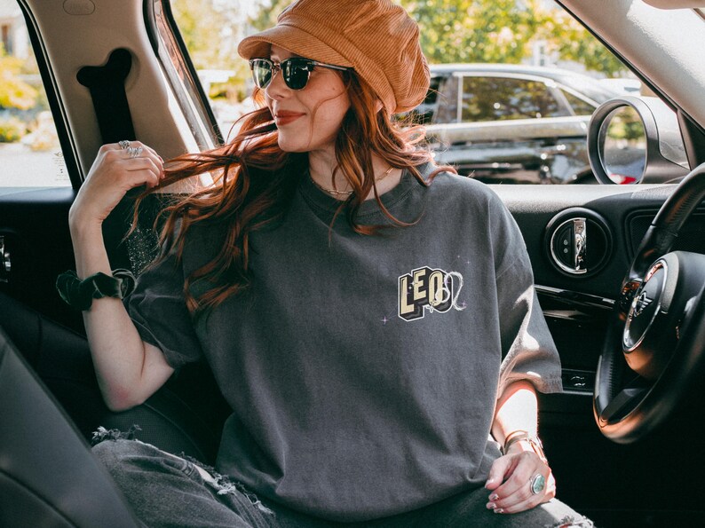 Puede incluir: Una mujer con una camiseta gris con la palabra "LEO" en letras amarillas en la parte delantera. Est&aacute; sentada en el asiento del conductor de un coche y lleva un sombrero de pana marr&oacute;n y gafas de sol.