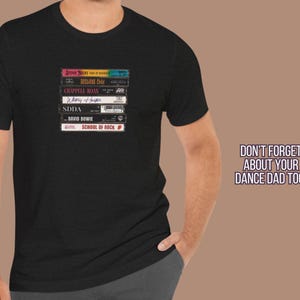 May include: A black t-shirt with a stack of cassette tapes featuring the band names "Journey", "Def Leppard", "Chappell Roan", "Whitney Houston", "SDDA", "David Bowie", and "School of Rock".