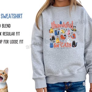 May include: A light gray kids' sweatshirt with the text "thankful for CATS" and various cartoon cat illustrations in multiple colors. The sweatshirt also has the text "KIDS SWEATSHIRT", "50/50 BLEND", "UNISEX REGULAR FIT", and "SIZE UP FOR LOOSE FIT".