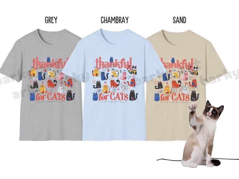 May include: Three t-shirts in grey, chambray, and sand colors, each with the text "thankful for CATS" and a colorful cat illustration. A cat is in the foreground, raising a paw.