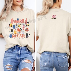 May include: Beige t-shirt with the words "thankful for CATS" and a colorful cat graphic. The back of the shirt has a pink cat graphic with the word "MEOW". The shirt is paired with blue jeans.