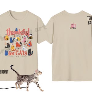 May include: Beige t-shirt with a colorful cat graphic. The front of the shirt says "thankful for CATS" in a playful font, with various cat illustrations. The back features a small pink cat and the word "MEW".