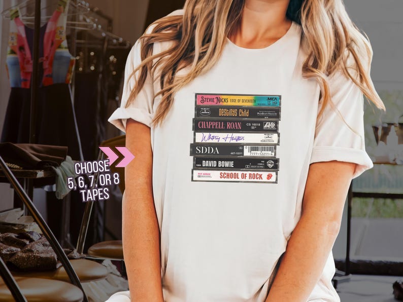 May include: A white t-shirt with a stack of cassette tapes printed on the front. The tapes are labeled with the names of various artists, including Stevie Nicks, Designs Child, Chappell Roan, Whitney Houston, SDDA, David Bowie, and School of Rock. The text "Choose 5, 6, 7, or 8 Tapes" is printed below the stack of tapes.
