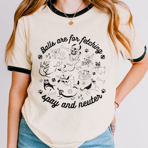 May include: A cream-colored t-shirt with a black ringer and a graphic of dogs playing fetch with the text "Balls are for fetching, spay and neuter".