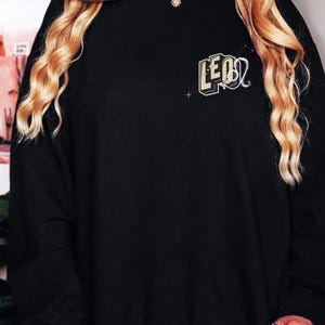 Puede incluir: Sudadera negra con un gr&aacute;fico dorado y negro del signo zodiacal Leo.