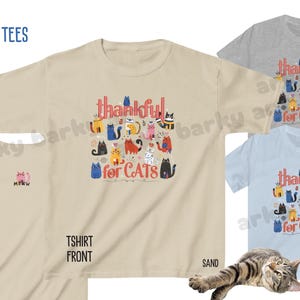 May include: A collection of kids' t-shirts in various colors, including sand, sport grey, and chambray. The front of the sand-colored shirt features the text "thankful for CATS" with colorful cat illustrations. The back of the sand shirt has a small "MEOW" graphic.