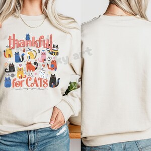 May include: Beige sweatshirt with the words "thankful for CATS" in a colorful, whimsical font. The design features various cartoon cats in different colors and patterns, creating a playful and cheerful aesthetic. The sweatshirt is a casual, comfortable style.