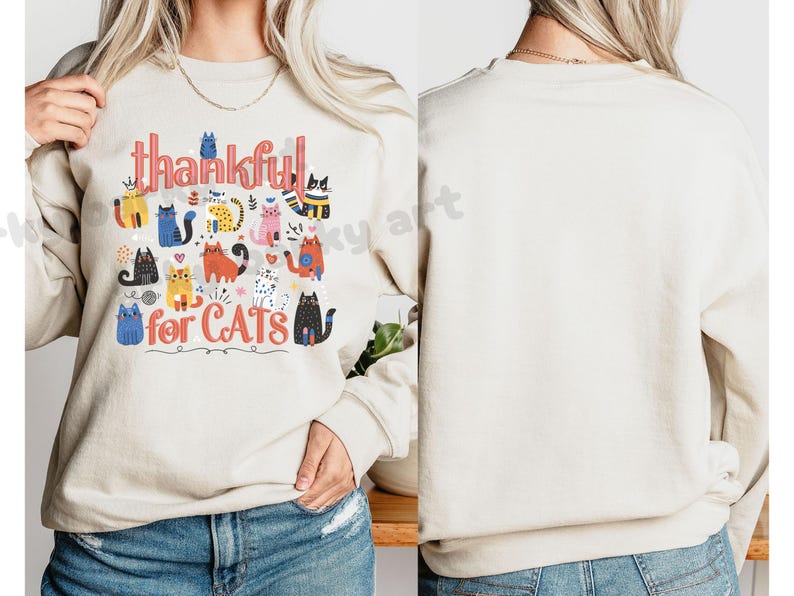 May include: Beige sweatshirt with the words "thankful for CATS" in a colorful, whimsical font. The design features various cartoon cats in different colors and patterns, creating a playful and cheerful aesthetic. The sweatshirt is a casual, comfortable style.