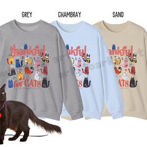 May include: Three sweatshirts in grey, chambray, and sand colors, each with a graphic of colorful cats and the text "thankful for CATS." A black cat with a purple collar and a red heart tag is in the foreground.