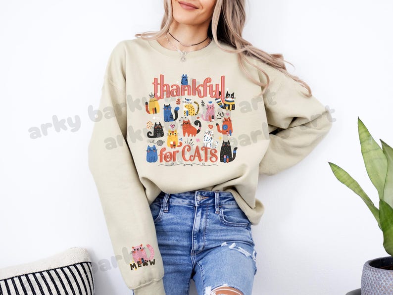 May include: Beige sweatshirt with the text "thankful for CATS" in red and black. The design features colorful cartoon cats. The sleeve has a pink cat graphic with the word "MEOW".