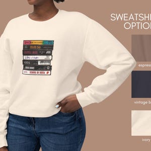 May include: A white sweatshirt with a stack of VHS tapes featuring the titles "Something for Joey", "Billy Idol", "Crowded House", "Mary J. Blige", "SODA", "David Bowie", and "School of Rock". The sweatshirt is available in espresso, vintage black, and ivory.