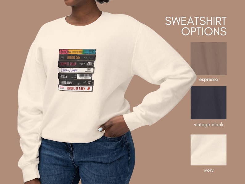May include: A white sweatshirt with a stack of VHS tapes featuring the titles "Something for Joey", "Billy Idol", "Crowded House", "Mary J. Blige", "SODA", "David Bowie", and "School of Rock". The sweatshirt is available in espresso, vintage black, and ivory.