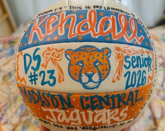May include: A white volleyball decorated with hand-painted orange and blue designs. It features the name Kendall, a jaguar mascot head, the text DS #23, senior 2026, Madison Central Jaguars, and the scripture Joshua 1:9 written in black ink.