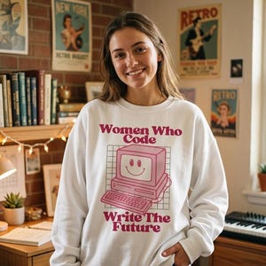 May include: White sweatshirt with the text "Women Who Code Write The Future" in a retro font. The graphic features a pink computer with a smiling face. The sweatshirt is a long-sleeved crew neck.