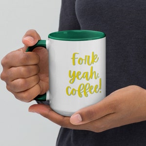 Puede incluir: Taza de café blanca con borde y asa verdes. La taza tiene el texto "Fork yeah. coffee!" en letras amarillas.