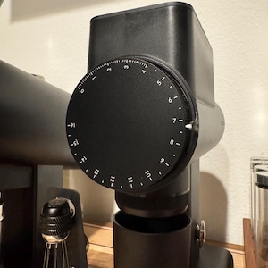 May include: A black and gray coffee grinder with a numbered dial. The dial has numbers from 0 to 16. The grinder has a cylindrical base and a rectangular top. The background is a light beige wall.