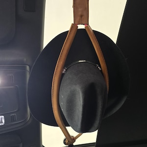 May include: A black hat suspended from a car's sunroof with a brown leather strap and suction cup. The hat is held in place by a leather strap with pink stitching.