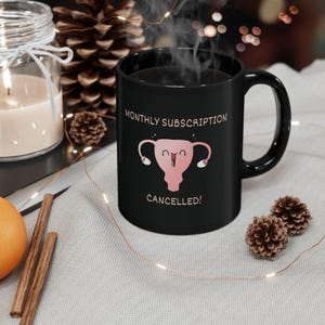 May include: Black coffee mug with a white outline and the text "MONTHLY SUBSCRIPTION CANCELLED!"  A cartoon uterus with a smiling face and pink color is in the center of the mug.