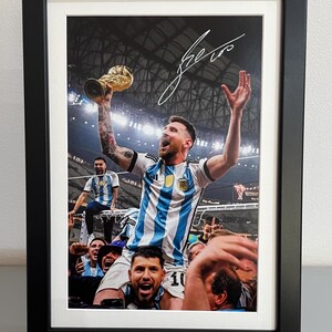 Könnte beinhalten: Gerahmter Druck eines Fußballspielers, der mit einer Trophäe feiert, umgeben von Teamkollegen. Der Spieler trägt ein blau-weiß gestreiftes Trikot. Der Druck befindet sich in einem schwarzen Rahmen mit weißem Passepartout.