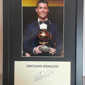 Może przedstawiać: Oprawione zdjęcie Cristiano Ronaldo trzymającego złoty puchar. Obraz jest umieszczony w czarnej ramie, z kremowym passe-partout. Poniżej zdjęcia znajduje się wydrukowany podpis i nazwisko "CRISTIANO RONALDO".