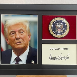 Puede incluir: Una foto enmarcada de Donald Trump con un marco negro y un sello dorado del Presidente de los Estados Unidos. El sello es rojo, blanco y azul. La foto está firmada por Donald Trump.