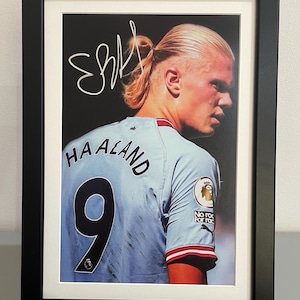 Könnte beinhalten: Gerahmter Druck eines Fußballspielers in einem hellblauen Trikot mit dem Namen "HAALAND" und der Nummer "9". Der Druck enthält eine weiße Unterschrift. Das Bild ist vor einem schwarzen Hintergrund und in Schwarz gerahmt.