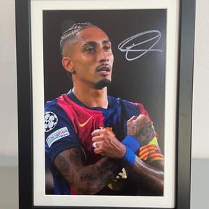 May include: Framed photograph of a soccer player in a team jersey, signed in silver ink. The player is wearing a blue and red jersey with a team logo. The photo is in a black frame and displayed on a wooden easel.