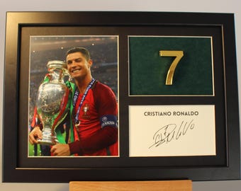May include: A framed photo of Cristiano Ronaldo holding the Euro 2016 trophy. The frame includes a gold number 7 and a signature. "CRISTIANO RONALDO" is printed below the signature.