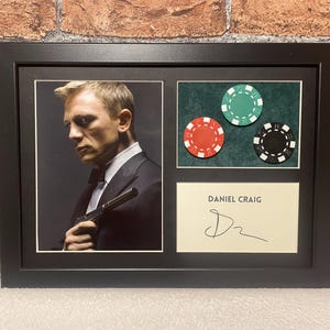 Puede incluir: Una foto enmarcada en negro de Daniel Craig, con un traje negro y sosteniendo un arma, con una firma y tres fichas de póquer en el fondo. Las fichas de póquer son verdes, rojas y negras.