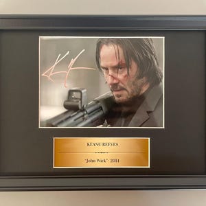 May include: Framed photograph of Keanu Reeves holding a gun, with a signature in the upper left corner. Below the photo is a gold plaque with the name "KEANU REEVES" and "John Wick" 2011.
