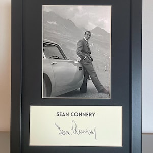 May include: Framed black and white photograph featuring Sean Connery next to a car, set against a mountain range. Beneath the photo is a card with "SEAN CONNERY" and his signature.