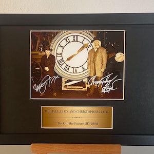 Può includere: Stampa incorniciata con Michael J. Fox e Christopher Lloyd da "Ritorno al futuro III". L'immagine mostra gli attori davanti a un grande orologio. La cornice nera circonda la stampa e una targa dorata in basso recita "Back to the Future III" - 1990.