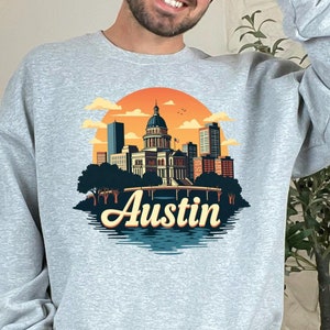 Op de afbeelding: Een grijs sweatshirt met een vintage-stijl illustratie van de skyline van Austin, inclusief het Texas State Capitol gebouw. De tekst "Austin" is gedrukt in een retro-lettertype onder de illustratie.