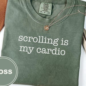 Könnte beinhalten: Moosgrünes T-Shirt mit dem weißen Text "scrolling is my cardio". Eine goldene Halskette mit Anhänger liegt auf dem Shirt. Ein brauner Hut und eine getrocknete Pflanze sind im Hintergrund.
