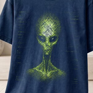 Könnte beinhalten: Ein marineblaues Kurzarm-T-Shirt mit einem Grafikdruck eines grünen Alien-Kopfes und Oberkörpers mit schraffierter Textur. Das T-Shirt hat einen Rundhalsausschnitt und wird auf einem Holzkleiderbügel präsentiert. Der Text ist sichtbar.