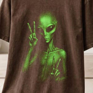 Könnte beinhalten: Ein braunes T-Shirt mit kurzen Ärmeln und einem Aufdruck eines grünen Aliens, das mit beiden Händen das Friedenszeichen zeigt. Das T-Shirt hat einen Rundhalsausschnitt und eine Vintage-Waschung. Der Text "OUTBOUND APPAREL COMPANY" ist oben links sichtbar.
