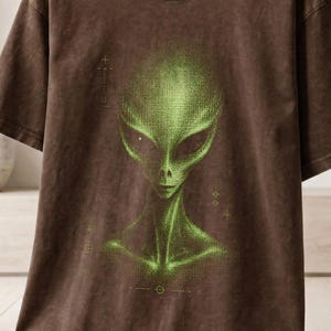May include: A brown short-sleeve t-shirt featuring a graphic of a green alien head and shoulders on the front. The shirt has a crew neckline and a vintage-style wash. A small circular badge in the bottom right corner displays the text ESPRESSO.