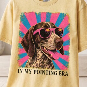 May include: A butter-colored short-sleeve t-shirt featuring a graphic of a brown and white dog wearing bright pink sunglasses. The dog is set against a blue and pink sunburst background with the text "IN MY POINTING ERA" printed below.