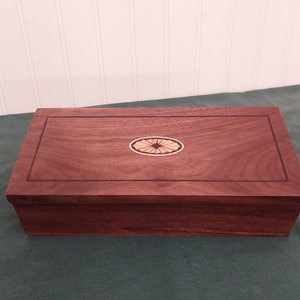May include: A wooden box with a dark brown finish and a decorative inlay in the center of the lid. The inlay is a light brown color and has a geometric design.