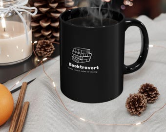 Puede incluir: Taza de cerámica negra con la palabra "Booktrovert" y una pila de libros en blanco. La taza tiene un asa curva y está llena de un líquido oscuro, posiblemente café o té. El texto debajo de la palabra "Booktrovert" dice "because I would rather be reading".