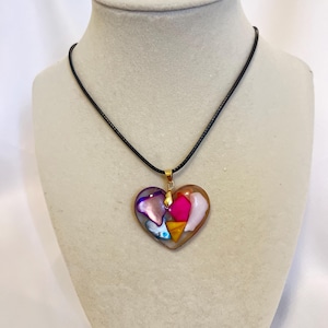 May include: A heart-shaped pendant necklace with a black cord. The pendant is made of clear resin with colorful pieces of abalone shell inside. The pendant is gold-toned.