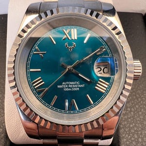 Puede incluir: Reloj de pulsera de acero inoxidable con esfera turquesa y agujas plateadas. El reloj presenta números romanos, una pantalla de fecha y el texto "AUTOMATIC WATER RESISTANT 100m:330ft". La correa del reloj también es de acero inoxidable.