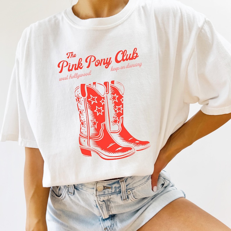 Op de afbeelding: Wit T-shirt met een rode grafische print van de tekst "The Pink Pony Club" met de woorden "west hollywood" en "keep on dancing" eronder. De grafiek bevat ook een paar rode cowboylaarzen met sterren.